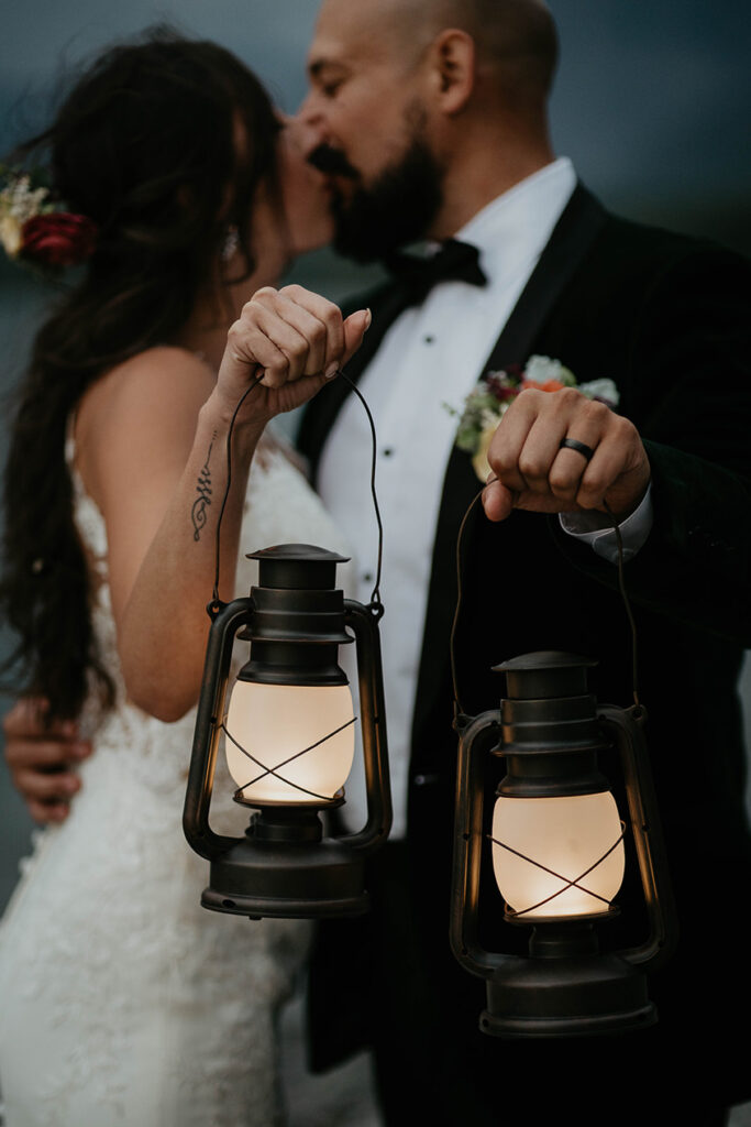 The couple holding lanterns standing on the lake shore with mountains around them and nighttime rolling in. 