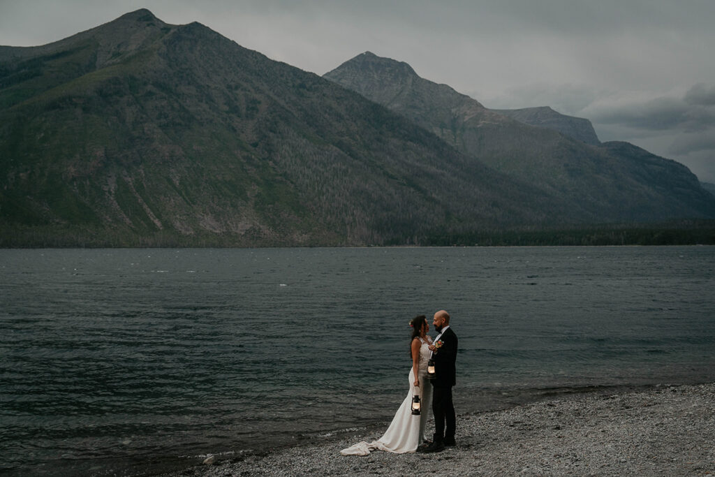 The couple holding lanterns standing on the lake shore with mountains around them and nighttime rolling in. 