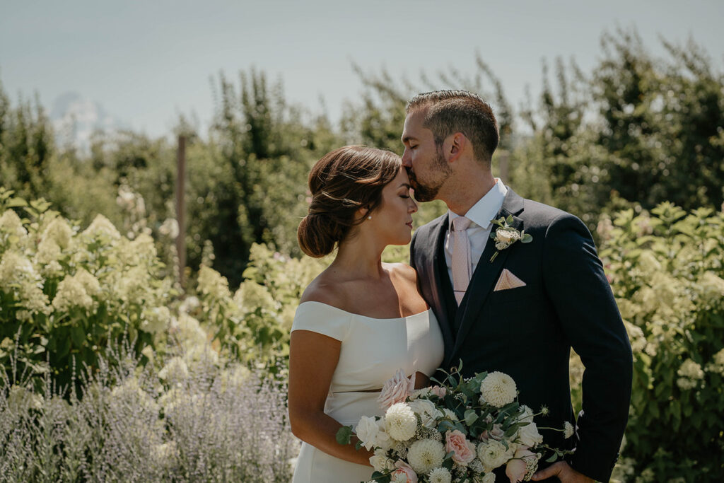 The groom kissing the bride's forehead with flowers and trees in the background. 