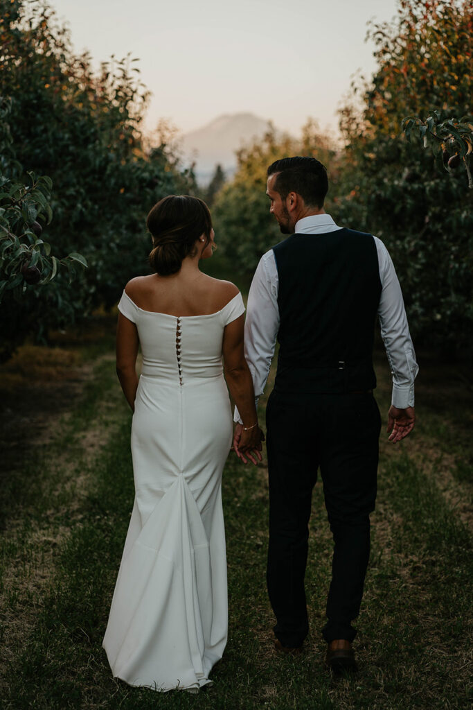 the bride and groom holding hands and walking through a grove of trees at The Orchard Hood River. 