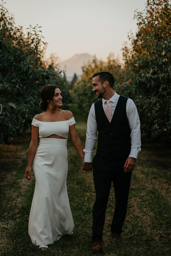 the bride and groom holding hands and walking through a grove of trees at The Orchard Hood River. 