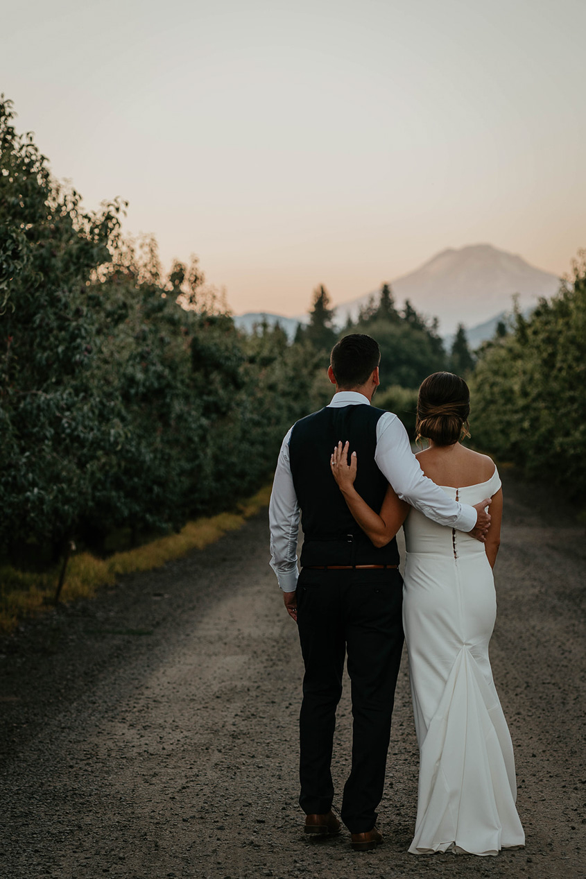 The bride and groom walking through a grove of trees side hugging each other. 