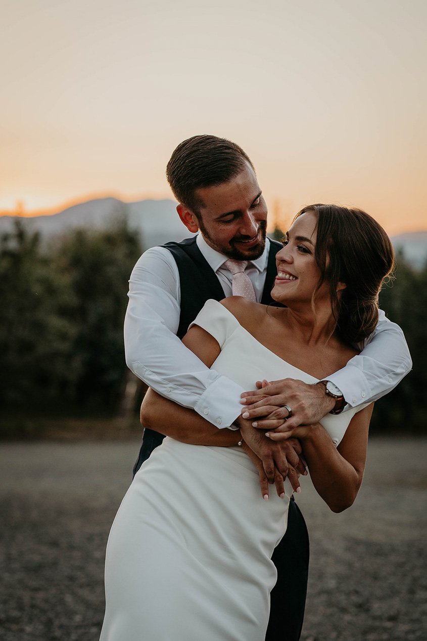 The groom hugging the bride and smiling at each other with a grove of trees behind them. 