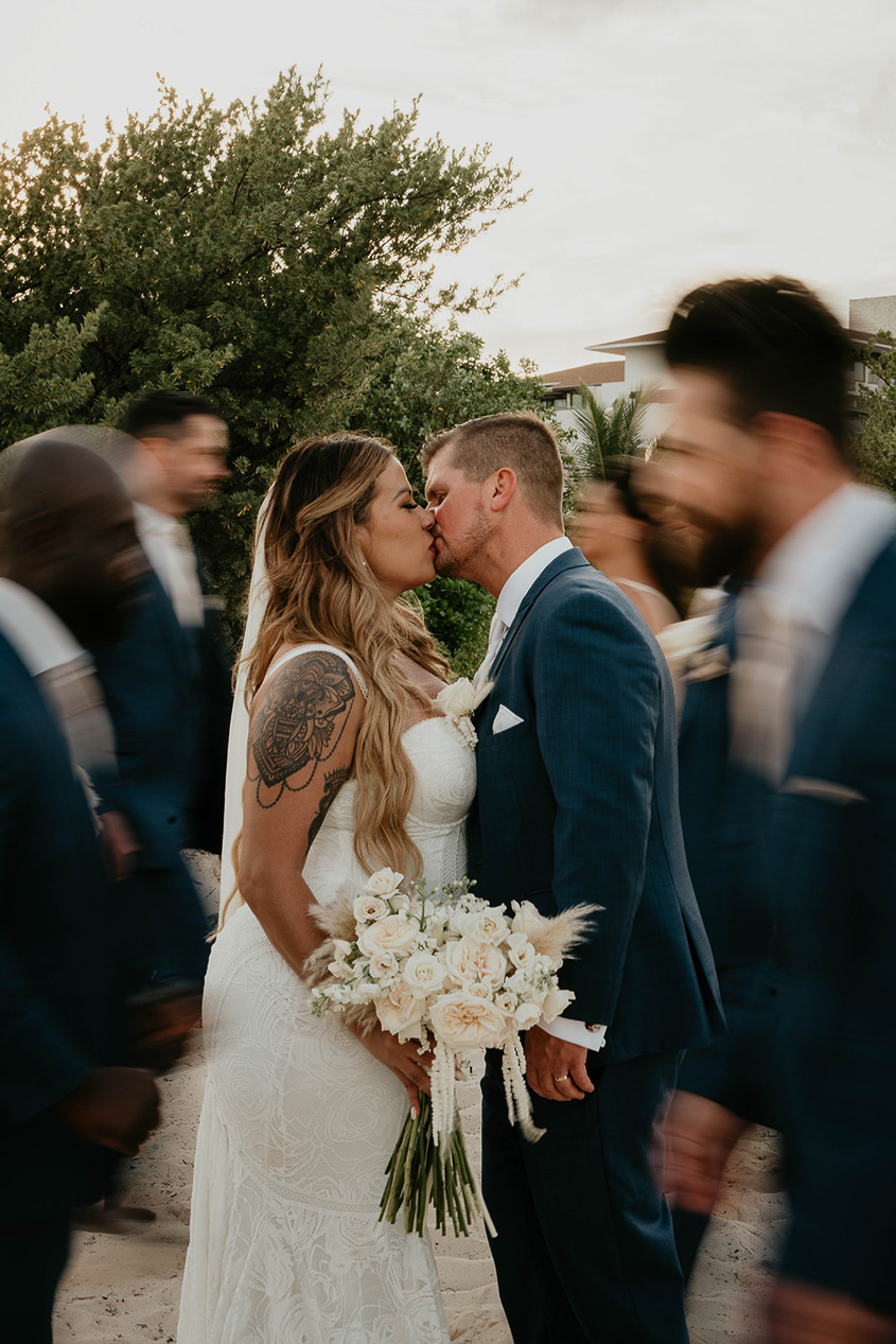 The bride and groom kissing with their wedding party blurred walking around them.