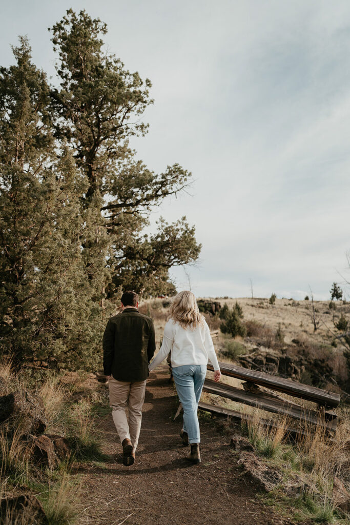 A couple holding hands walking away from the camera on a trail. 