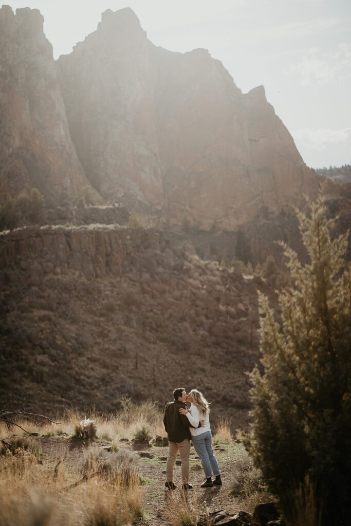 The couple kissing with big rocks in the distance during their Smith Rock engagement session. 