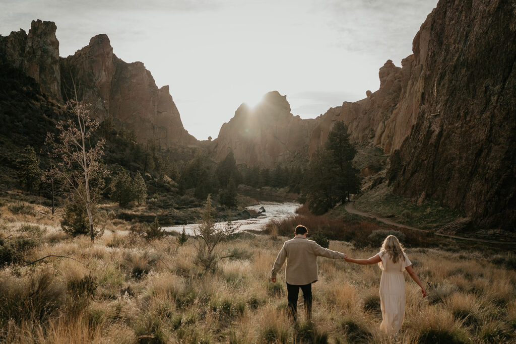 The couple walking through a field at sunset during their Smith Rock engagement session. 