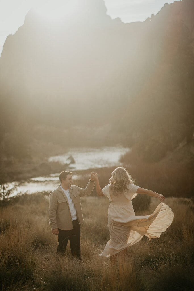 The couple dancing in a field with large spires and a river in the background during their Smith Rock engagement session. 