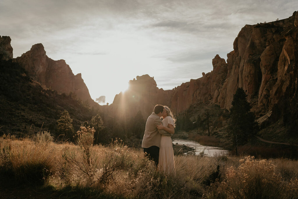 a couple kissing at sunset with larg spires and a river in the background during their Smith Rock engagement session. 