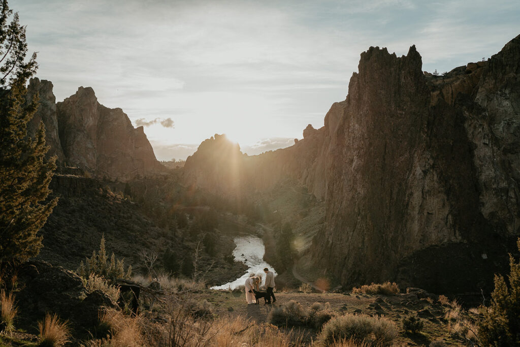 A couple walking through a field with their dog and featuring a river and spires in the background during their Smith Rock engagement session. 