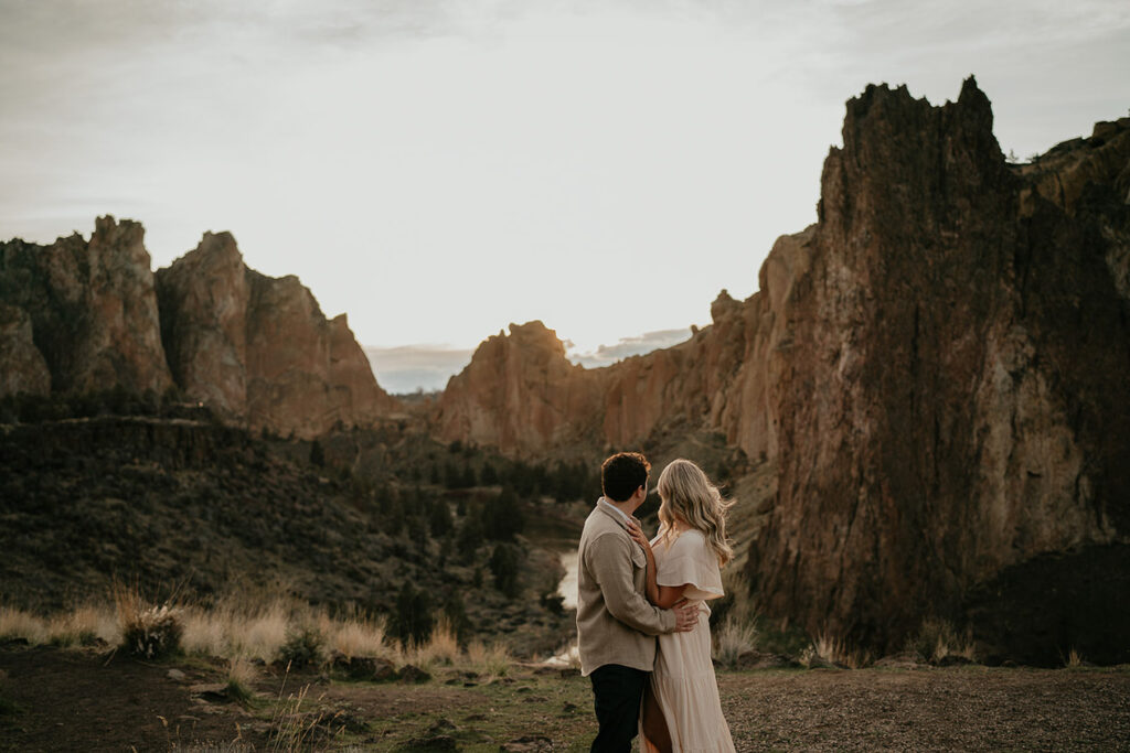 The couple holding each other with a river and cliffs in the background during their Smith Rock engagement session. 