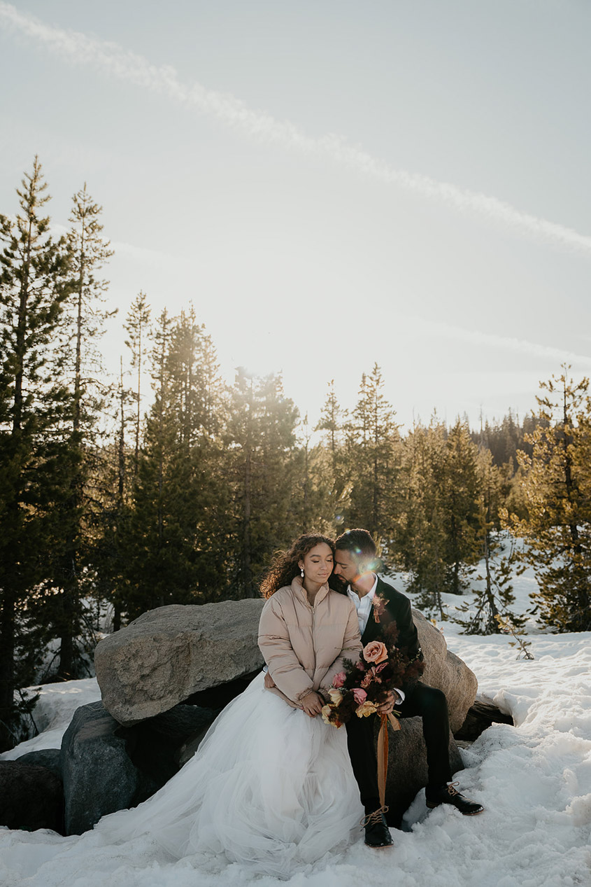 The newlyweds sitting on a rock in the snow, with pine trees in the background. 