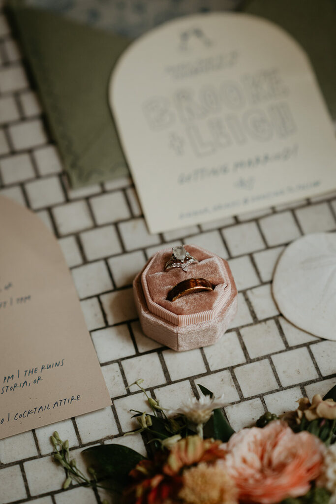 A close up of wedding rings, as well as a wedding invite, flower, and sea shell. 