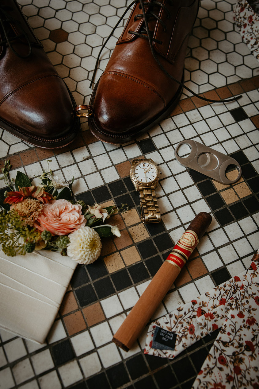 A cigar, tie, flowers, watch, cigar cutter, boots, and a wedding ring on a tile floor. 