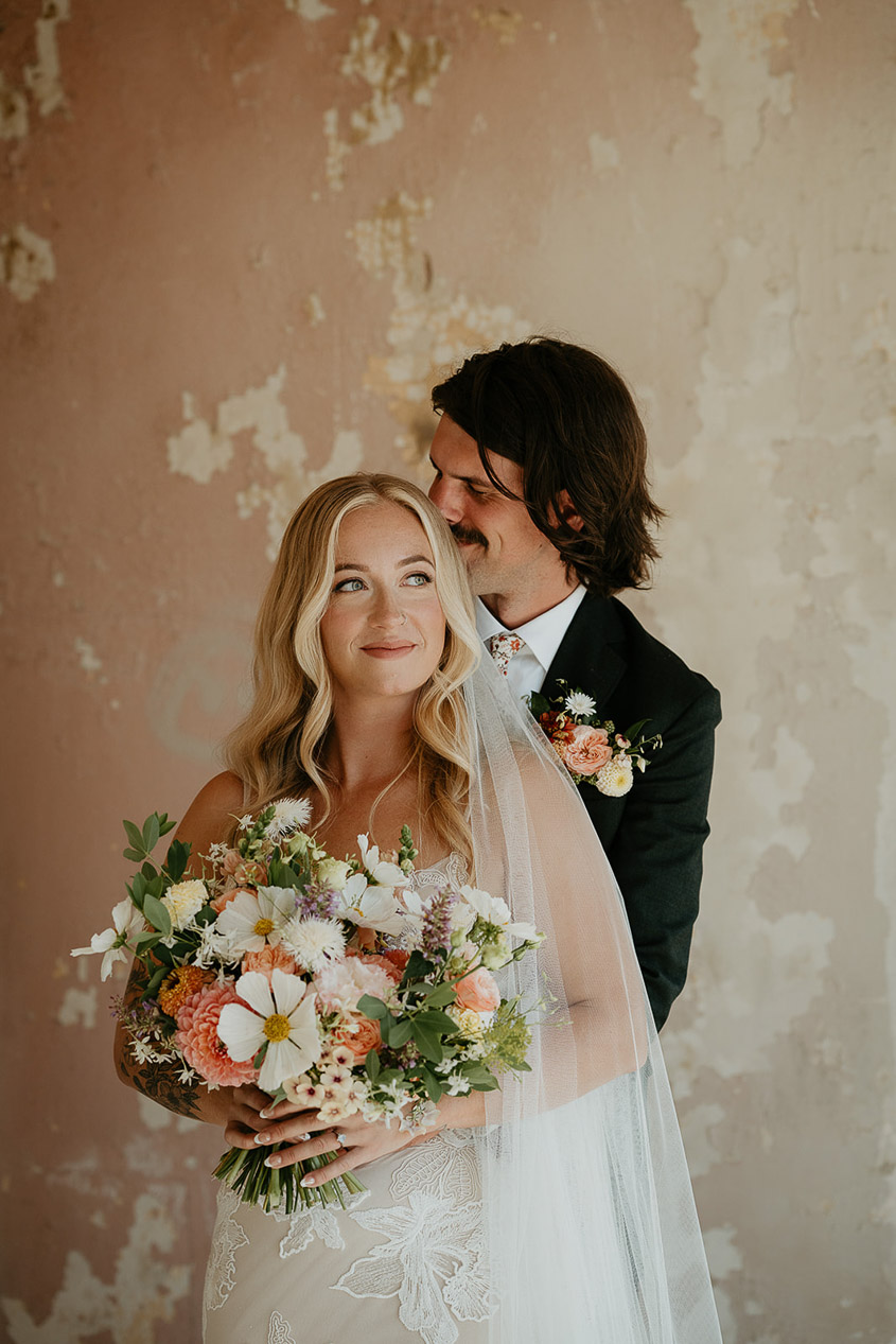 The groom hugging his wife while she holds a bouquet of flowers. 