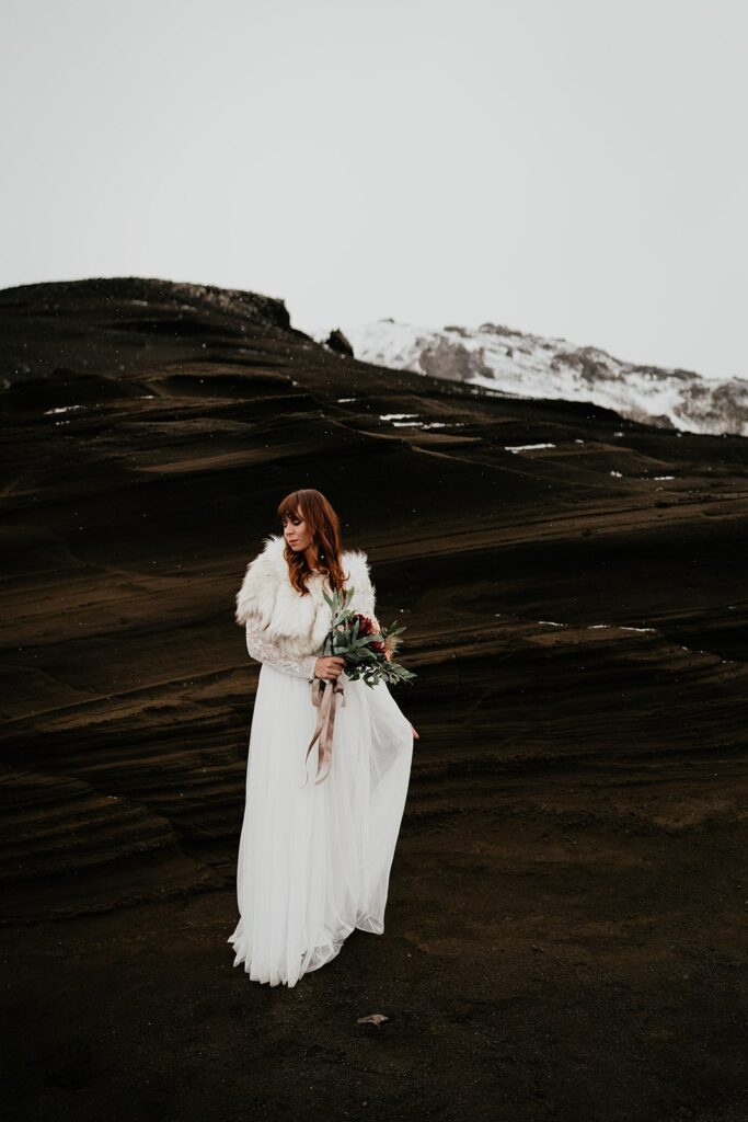 Bride holds the train of her white wedding dress while standing on a black sand beach at her elopement in Iceland 