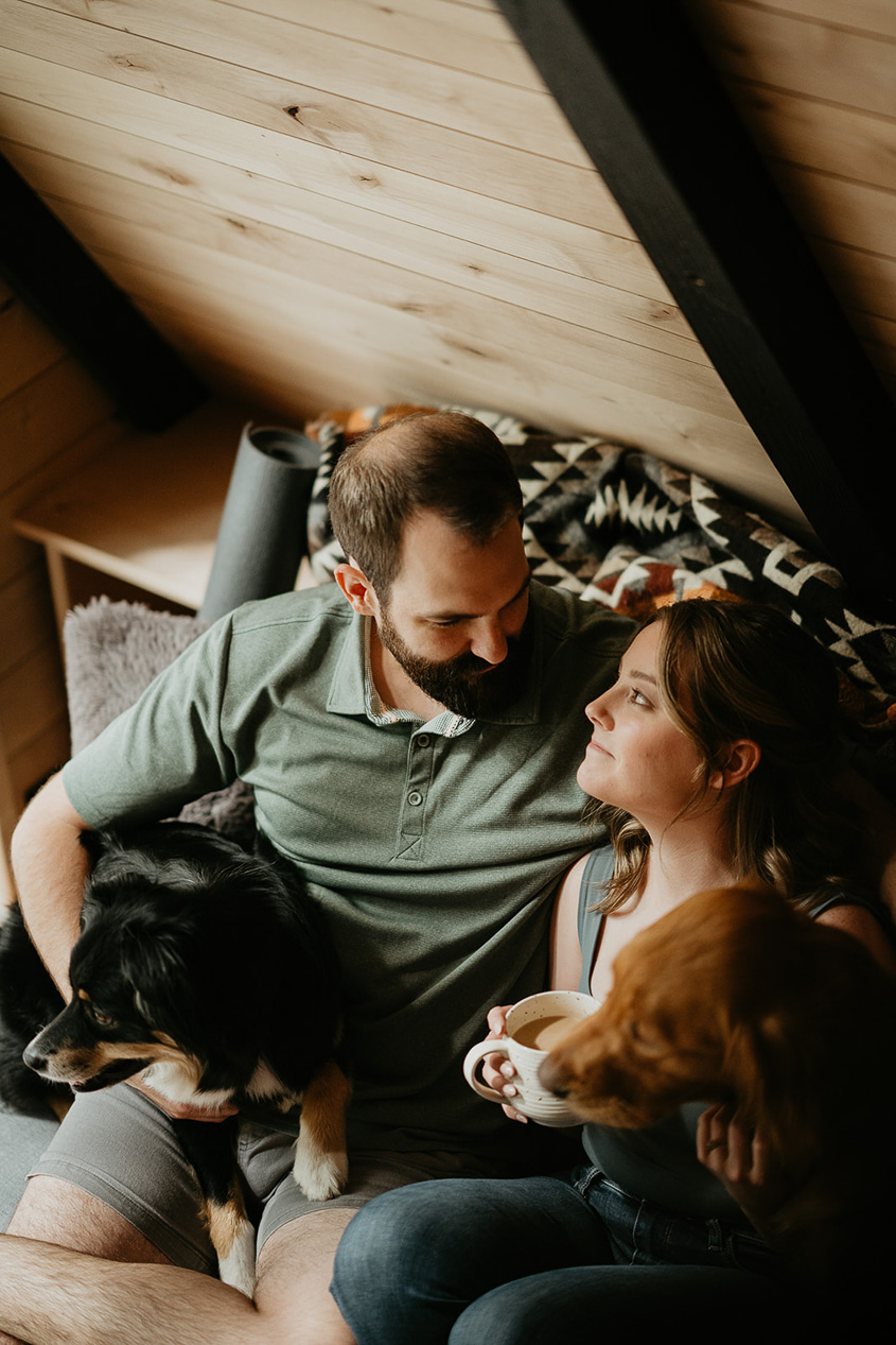 a couple sitting on a couch with their dogs in a cabin, happy about how inexpensive the cost is to elope.
