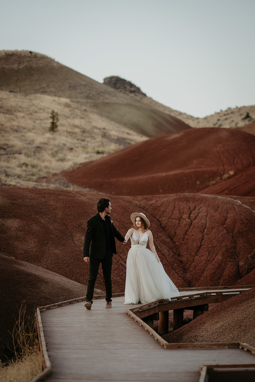 The groom and bride walking on a boardwalk through the painted hills in Oregon during their elopement.