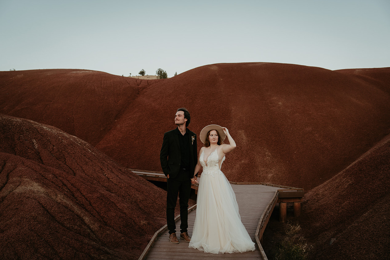 The newlyweds posing on a boardwalk with red mounds behind them during their painted hills elopement.
