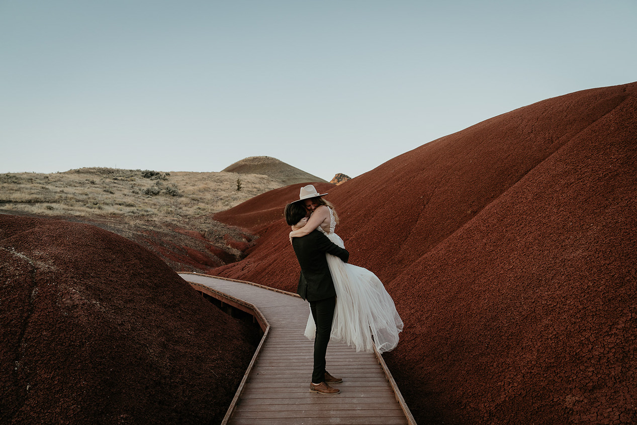 the newlyweds hugging on a boardwalk with red hills around them during their painted hills elopement.