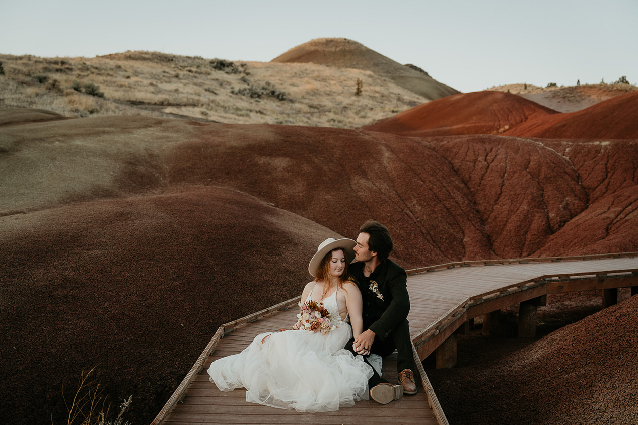 newlyweds holding each other sitting on a boardwalk through red hills.