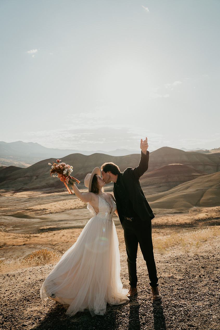 newlyweds kissing at sunset with a golden field and hills in the background.