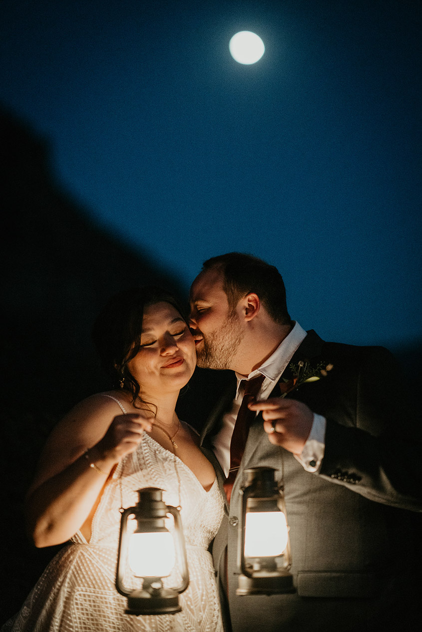 the groom kissing his bride under the moonlight, while they both are holding lanterns, during their wind mountain ranch wedding.