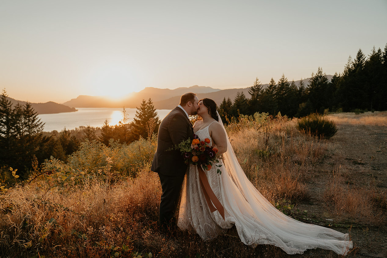the newlyweds kissing at sunset with a lake and mountains in the background during their wind mountain ranch wedding.