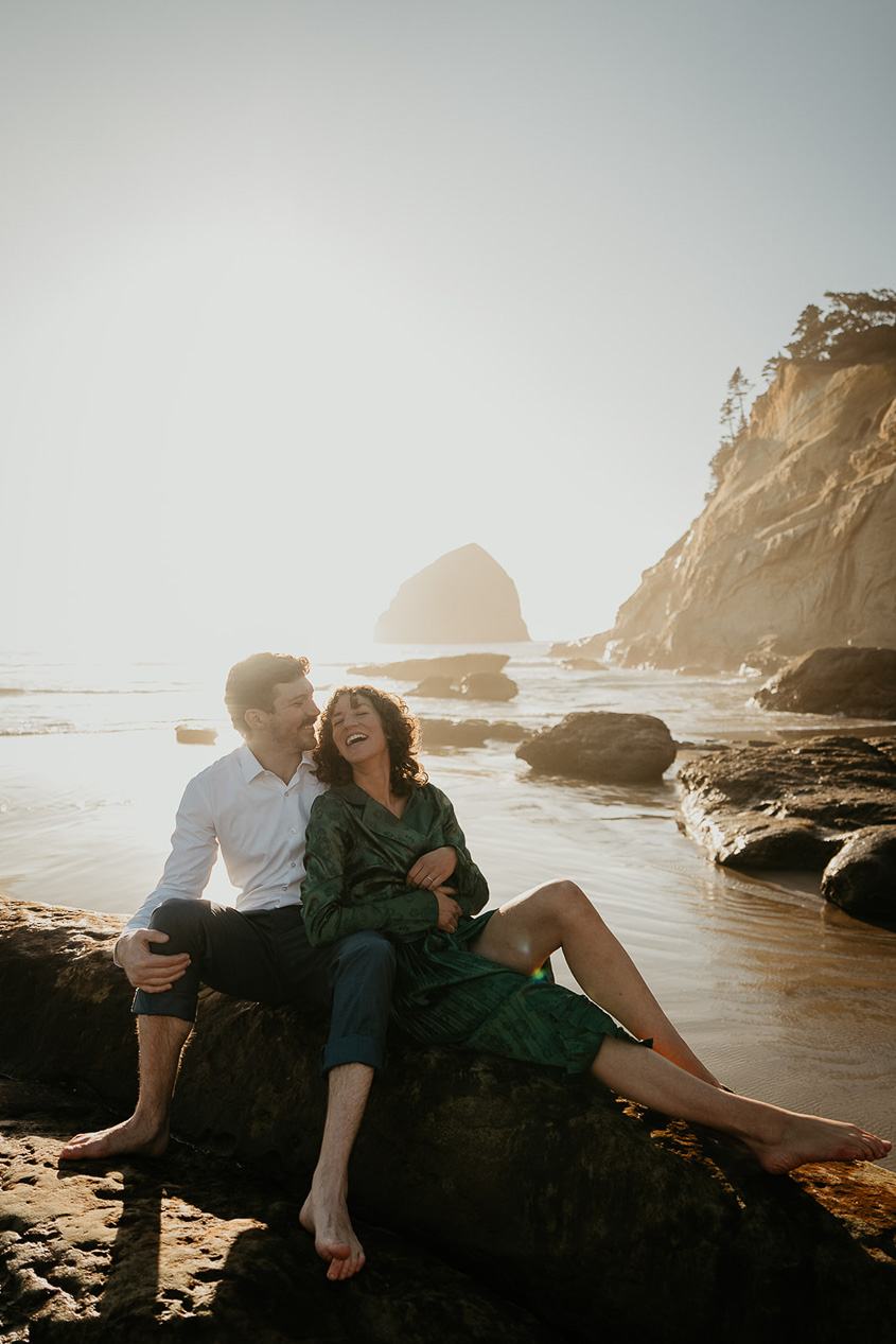 a couple smiling and sitting on a log along the Oregon coast.