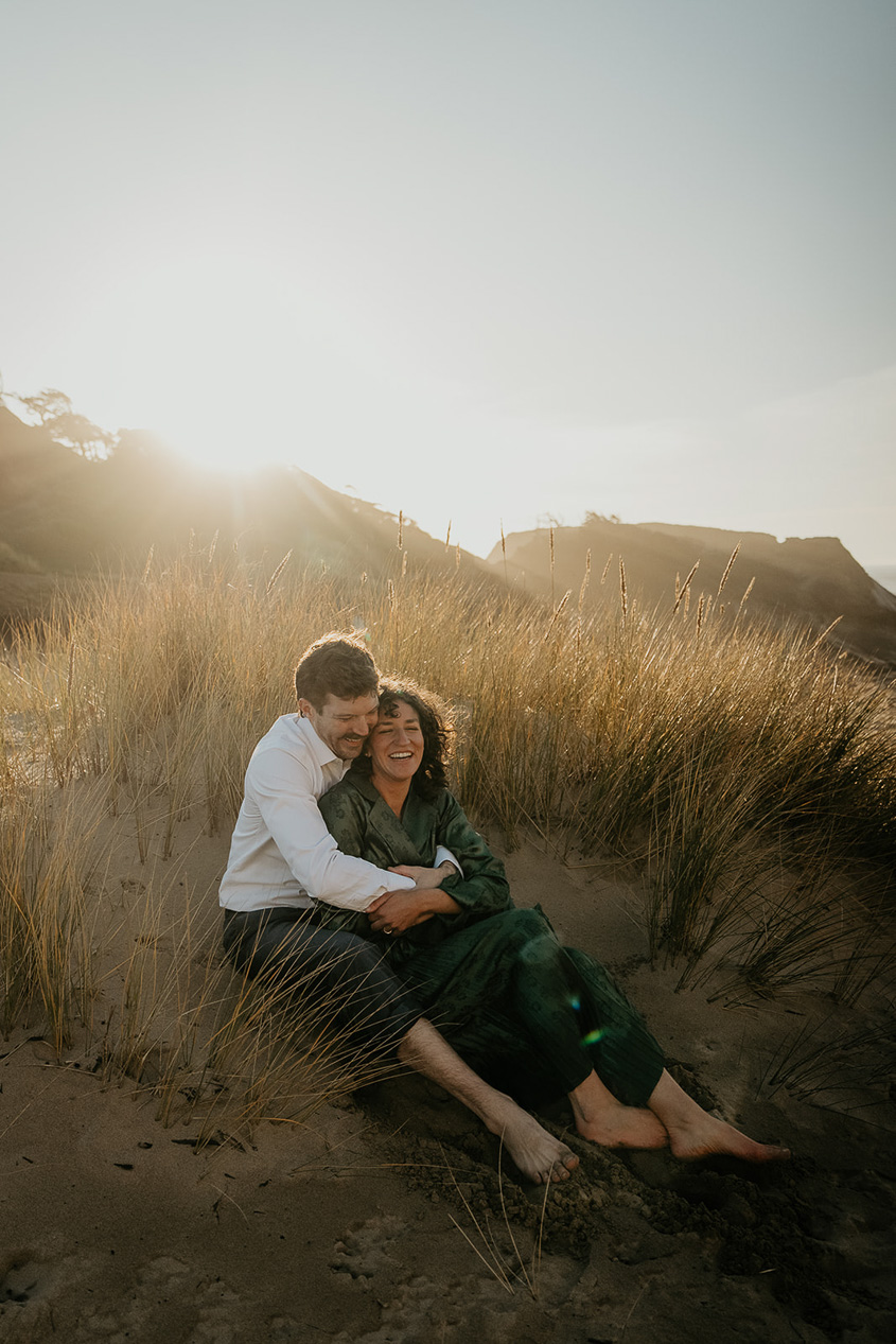 a couple smiling and sitting on the sand, surrounded by grasses.