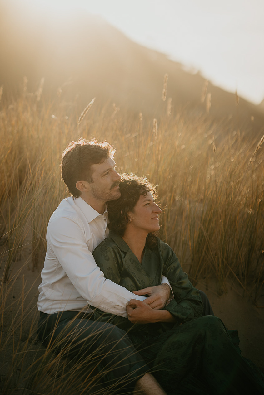 a couple hugging each other sitting in a grassy field during their cape kiwanda engagement.