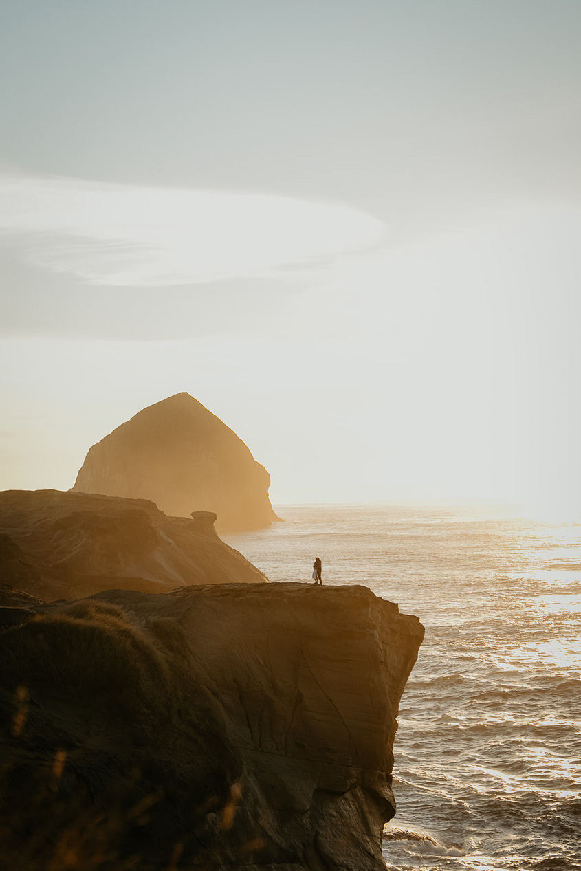 a couple holding each other standing on a cliff at sunset during their Cape Kiwanda Engagement.