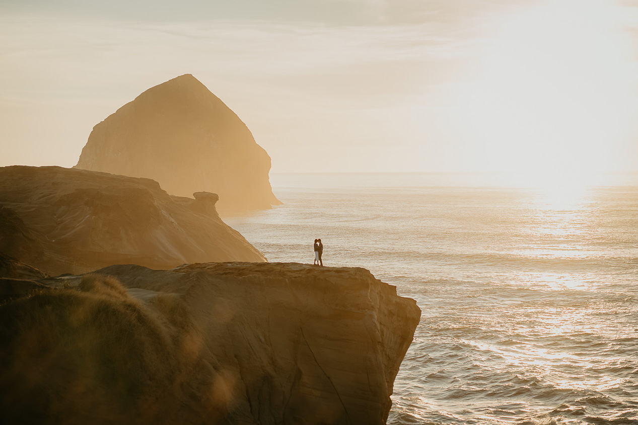 a couple holding each other with the coastline and a sea stack in the background during their Cape Kiwanda Engagement.