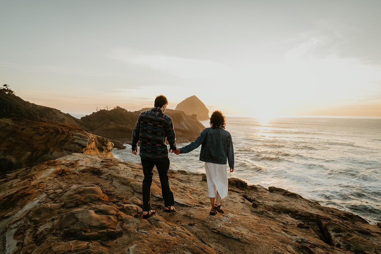 a couple holding hands walking along the cliffside above the ocean during their Cape Kiwanda Engagement.