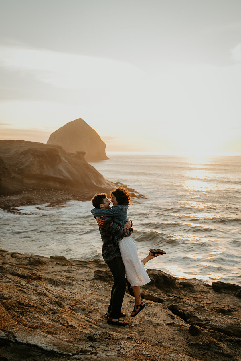 a couple hugging on a cliffside with the Oregon coastline in the background at sunset during their Cape Kiwanda Engagement.