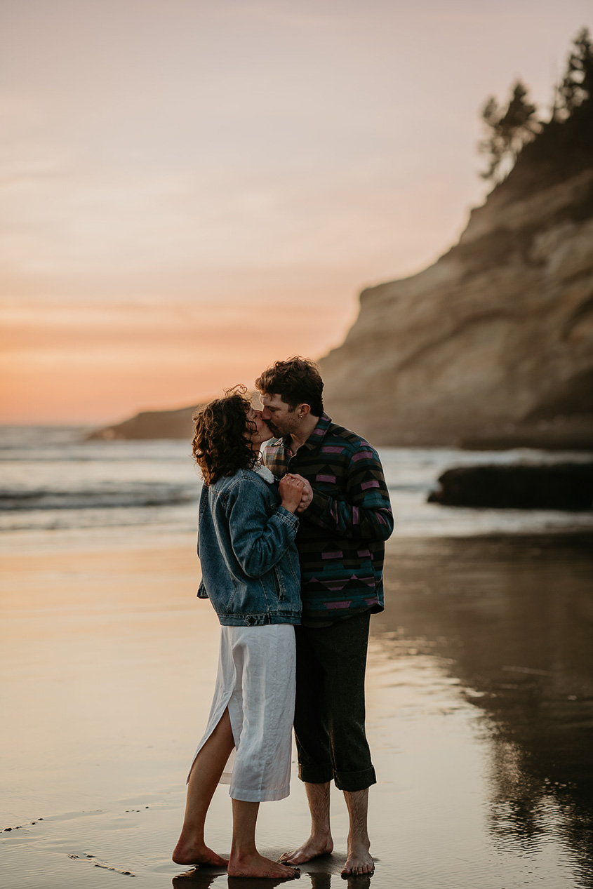 a couple kissing while standing on the sand during their Cape Kiwanda Engagement.