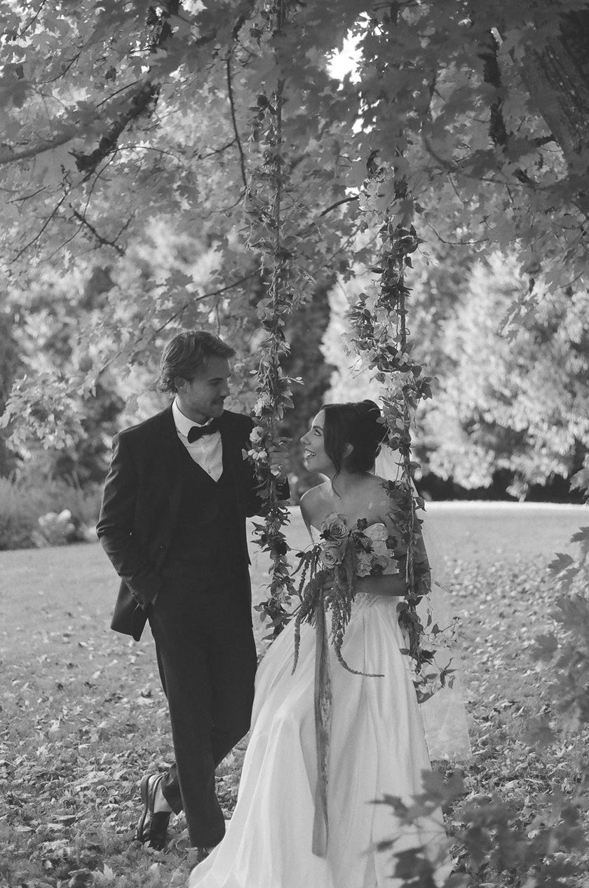 the groom standing next to his bride, who's sitting on a swing.