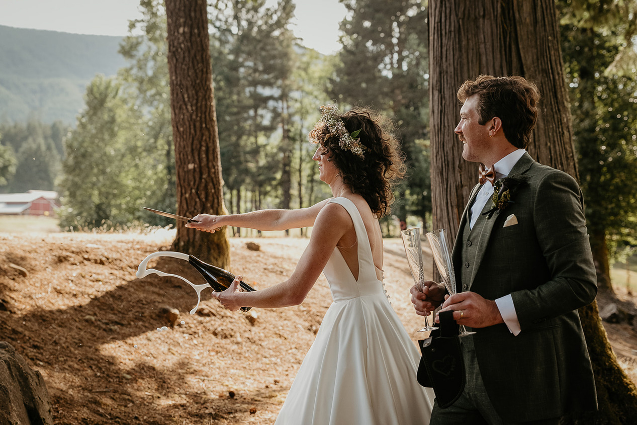 the bride sabering a champagne bottle while the groom watches behind her.