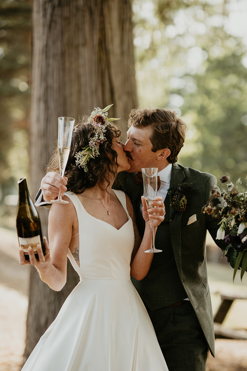the newlyweds holding up a bottle and glasses filled with Champagne while kissing during their rustic barn wedding.