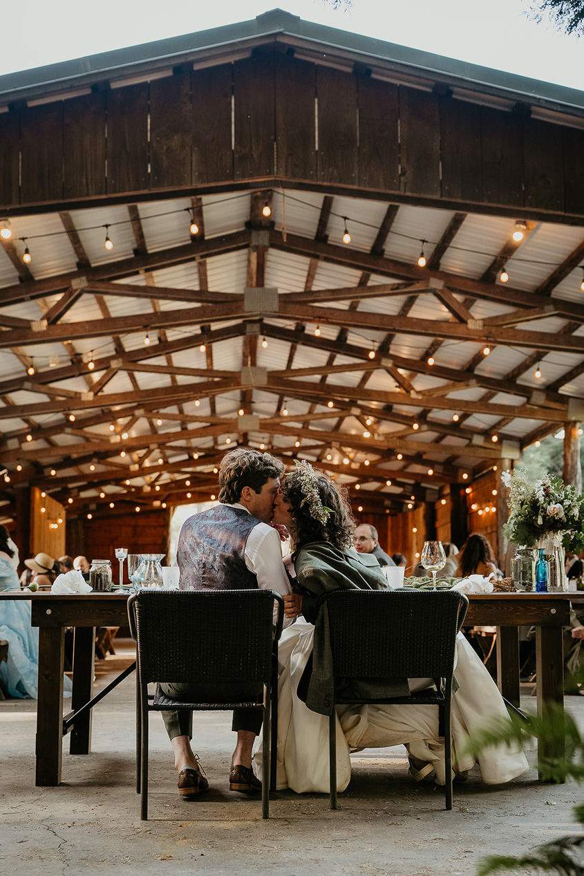 the newlyweds kissing at their dinner table during their rustic barn wedding.