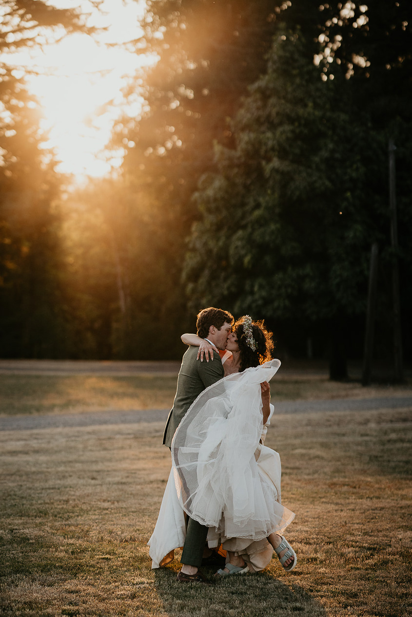 newlyweds kissing in a grassy field at sunset.