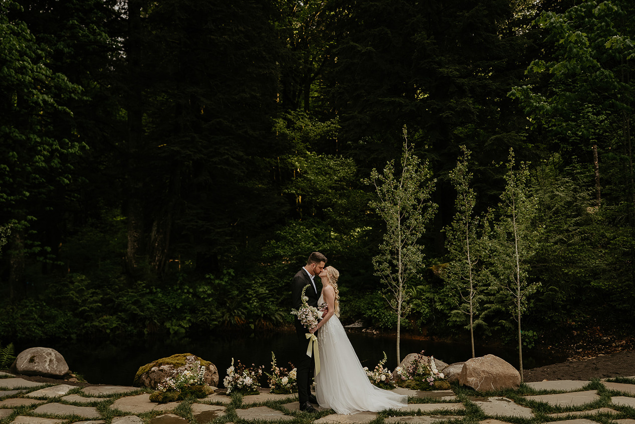 newlyweds kissing at the Highland Farms wedding venue with a pond and forests in the background.