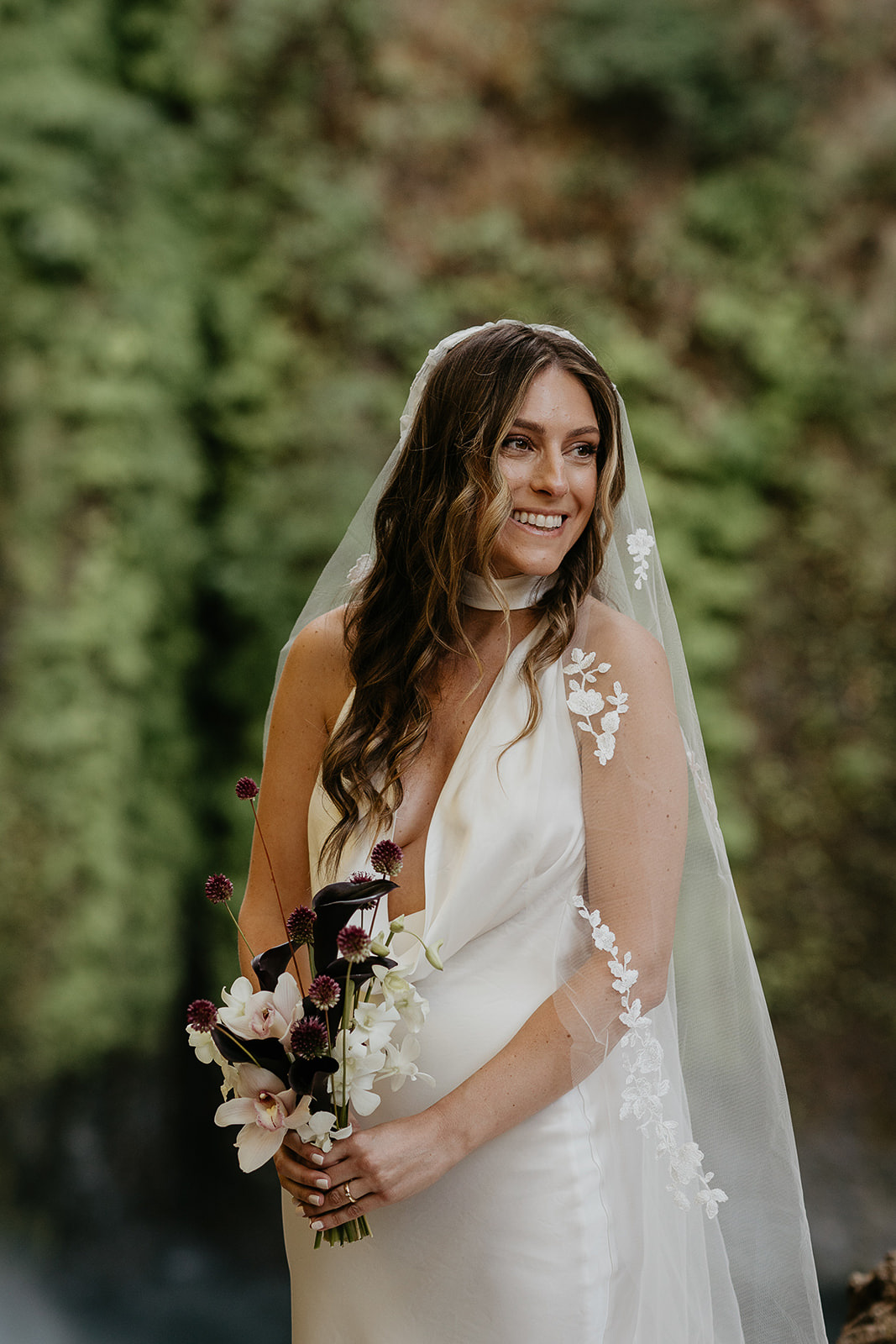 the bride holding a bouquet of flowers smiling in front of a mossy backgorund.