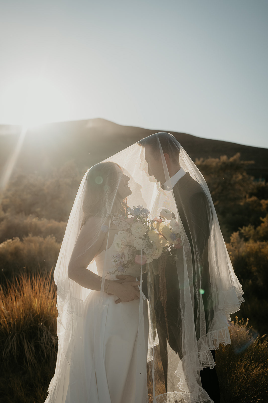newlyweds admiring each other under the bride's veil at sunset.