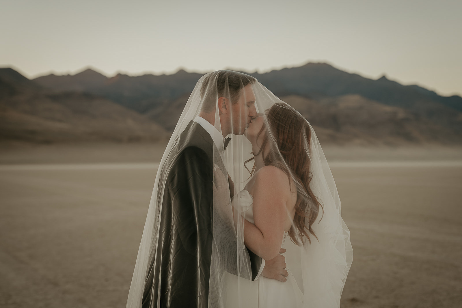 newlyweds kissing under the bride's veil on a desert playa at sunset.