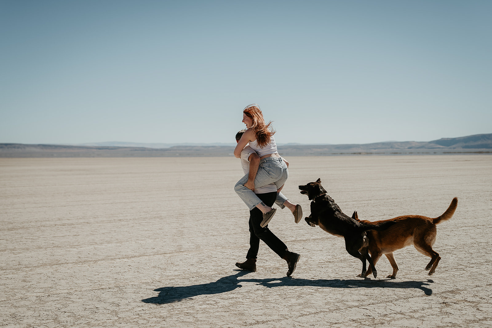 the husband carrying his wife on his back while their dogs playfully chase after them.
