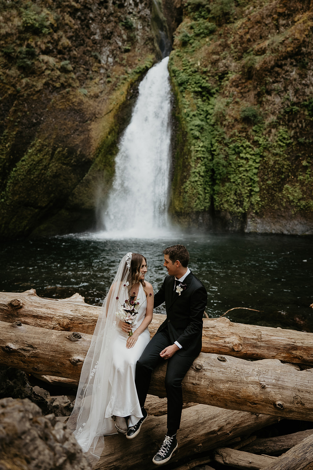 newlyweds sitting on a log by a waterfall during their elopement.