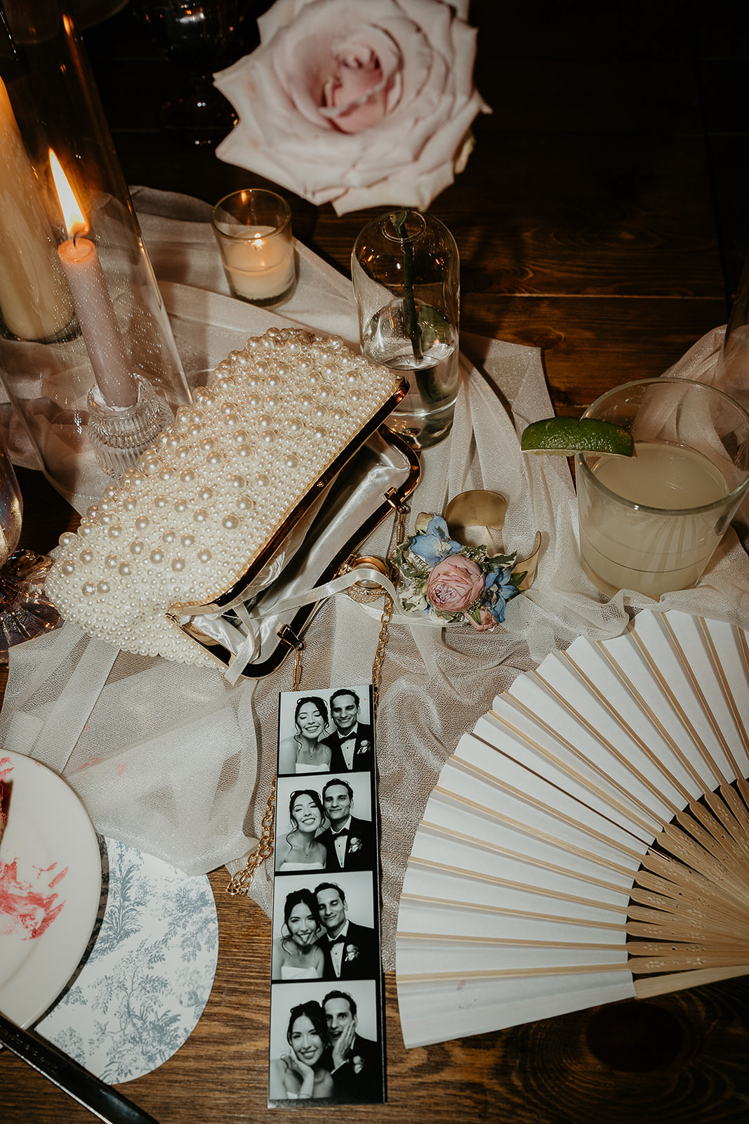 a table full of wedding memories, including a fan, rose, a drink, photo booth photos, and a clutch.