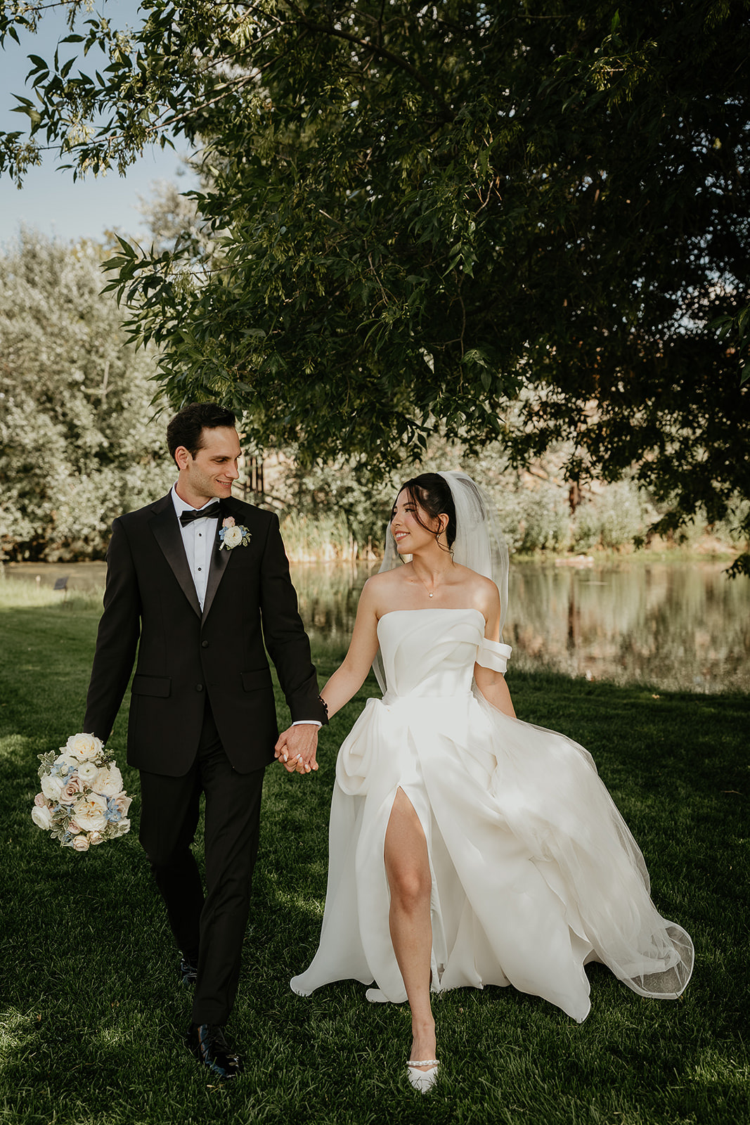 newlyweds holding hands under a tree with a pond in the background during their Brasada Ranch wedding.