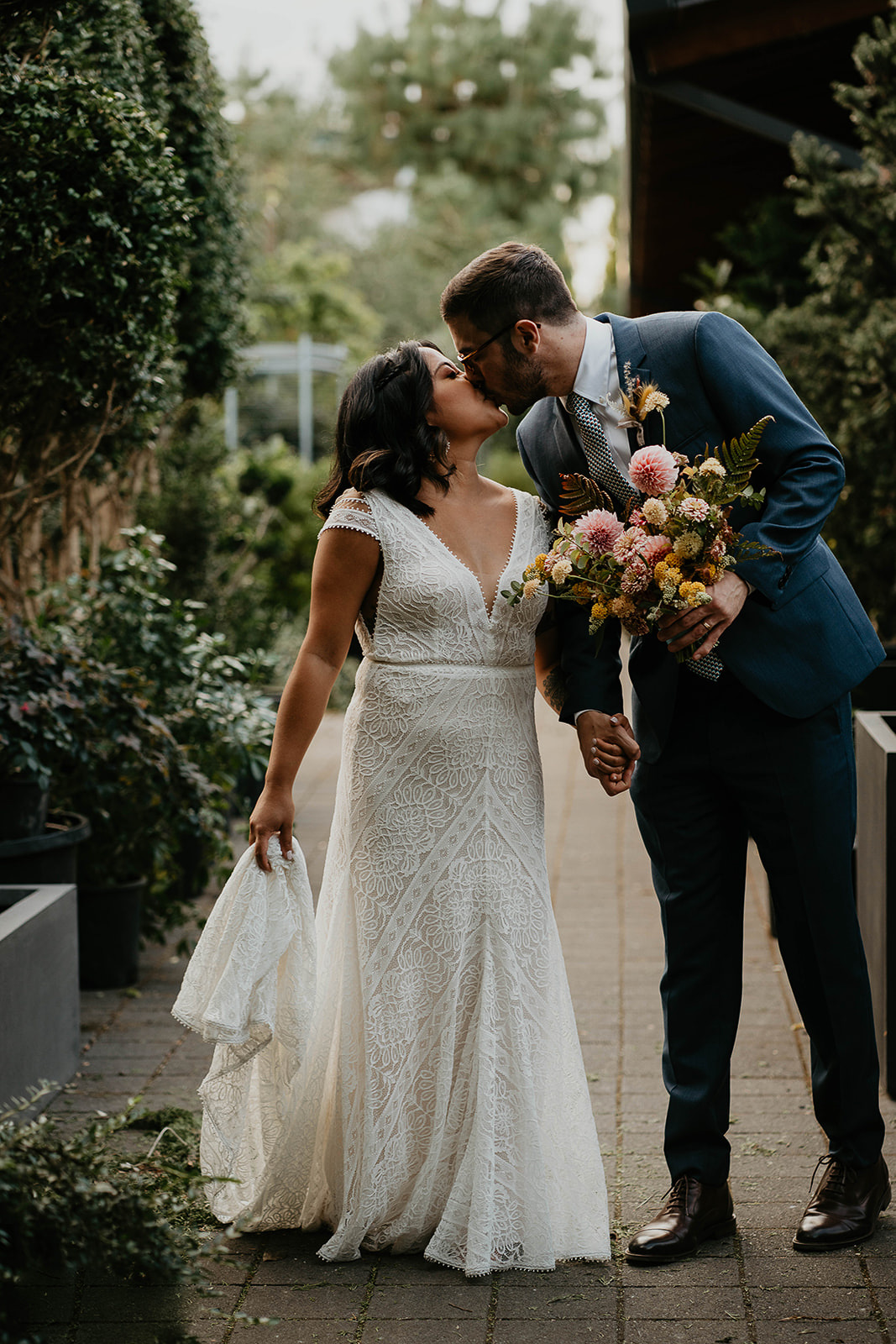 newlyweds kissing on a brick path by plants.